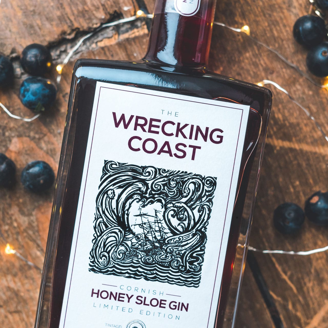 A photograph of Thew Wrecking Coast Distillery Honey Sloe Gin bottle on wooden table top surrounded by sloes